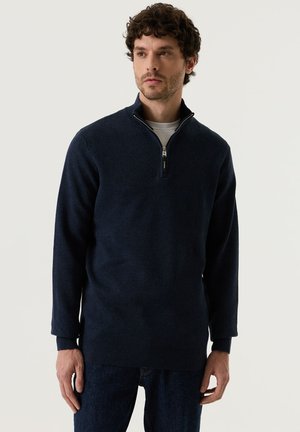 Mann steht vor schlichtem Hintergrund, trägt einen marineblauen Viertelreißverschluss-Pullover, ein weißes Unterhemd und dunkle Jeans und schaut leicht zur Seite.