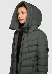 Veste rembourrée à capuche de couleur vert foncé, avec une fermeture éclair à l'avant, des cordons de serrage ajustables et un tissu texturé avec des motifs matelassés horizontaux.