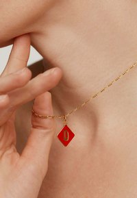 Collar de cadena de oro con un colgante rojo en forma de diamante que presenta la letra "O" en oro. El colgante tiene un acabado brillante y una textura suave.