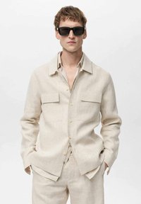 Bežena lanena srajca s ovratnikom, dvema prsnih žepoma in gumbi, v kombinaciji z usklajenimi hlačami. Manekenka nosi črne sončna očala.