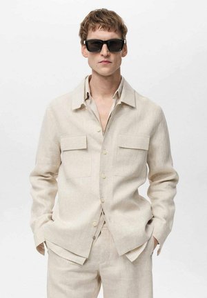 Camicia di lino beige con colletto, due tasche sul petto e bottoni, abbinata a pantaloni coordinati. Modello che indossa occhiali da sole neri.