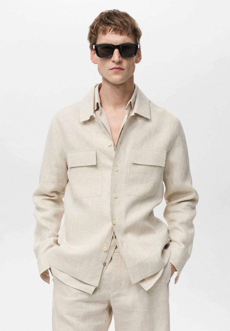 Bežena lanena srajca s ovratnikom, dvema prsnih žepoma in gumbi, v kombinaciji z usklajenimi hlačami. Manekenka nosi črne sončna očala.