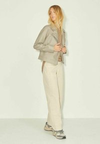 Giacca in pelle grigia chiara con tasche con zip, abbinata a una maglietta beige e pantaloni color beige. Modella che indossa sneaker grigie e beige.