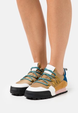 Pieds portant des baskets beige, blanches, noires et bleues avec des lacets turquoise, debout sur une surface blanche.