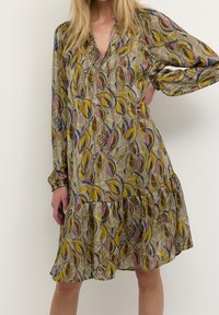 Mønstret kjole med en blanding af gule, brune og blå bladdesigns. Let stof med lagdelt kant og lange ærmer. V-hals detaljer.