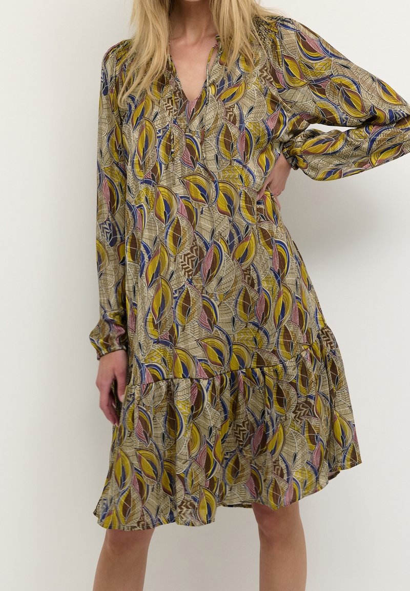 Mønstret kjole med en blanding af gule, brune og blå bladdesigns. Let stof med lagdelt kant og lange ærmer. V-hals detaljer.