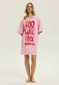 Młoda kobieta stojąca boso, ubrana w luźną różową sukienkę z czerwonym napisem "TOO CUTE TO BE alone" na jasnym, jednolitym tle.