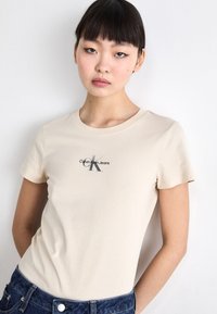Bežové bavlnené tričko s okrúhlym výstrihom, krátkymi rukávmi a potlačou loga "Calvin Klein Jeans" so štylizovaným 'CK' v sivej farbe.