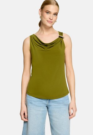 Jeune femme aux cheveux clairs portant un haut sans manches vert olive avec un col drapé et un jean bleu clair sur fond blanc.