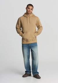 Felpa con cappuccio beige con tasca frontale, cordini e polsini a costine, abbinata a jeans in denim azzurro chiaro e scarpe scure.