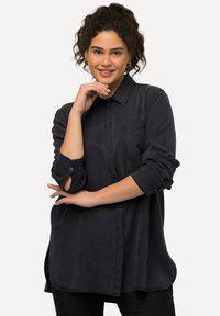 Camicia button-up blu scuro con maniche lunghe, taschino singolo sul petto, orlo curvo e texture del tessuto morbida. Presenta maniche arrotolate.