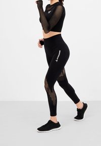 Schwarzes Activewear-Set bestehend aus einem langärmligen Mesh-Crop-Top und enganliegenden Leggings mit Mesh-Einsätzen. Die Leggings verfügen über ein Logo-Detail "SQUATPROOF".