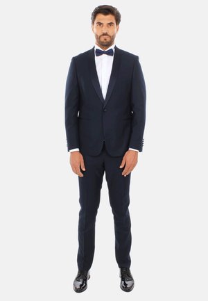 Tuxedo navy con rever in raso, camicia bianca e papillon nero. Pantaloni aderenti e scarpe eleganti nere lucide completano l'outfit.