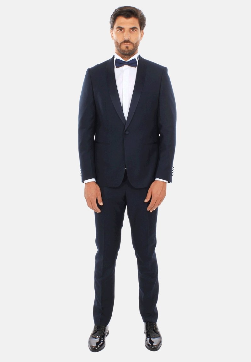 Tuxedo navy con rever in raso, camicia bianca e papillon nero. Pantaloni aderenti e scarpe eleganti nere lucide completano l'outfit.