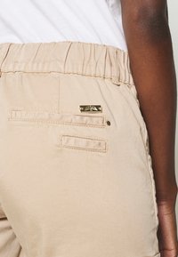 Pantalon beige à taille élastique avec des poches arrière, dont une ornée d'un détail en métal doré. Le tissu semble léger avec une texture lisse.