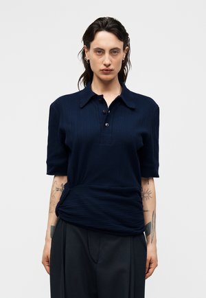 Femme aux cheveux foncés portant une chemise côtelée bleu marine à manches courtes avec col et un pantalon noir, montrant des tatouages sur les deux avant-bras sur un fond uni.