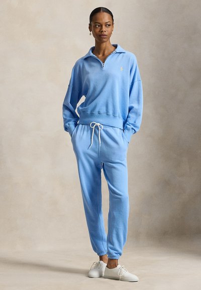Γυναίκα με ανοιχτό μπλε φούτερ με φερμουάρ και ταιριαστό παντελόνι φόρμας, με λευκά αθλητικά παπούτσια, στέκεται με τα χέρια στις τσέπες μπροστά σε ουδέτερο φόντο.