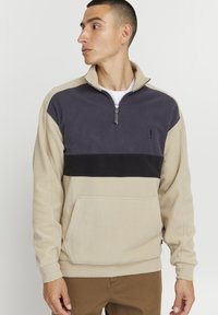 Sudadera de forro polar que presenta un cuerpo beige, cuello gris oscuro y una franja horizontal negra. Con bolsillo frontal y cierre de cremallera de cuarto. Textura suave.