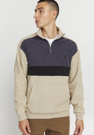 Fleecepullover med en beige kropp, mörkgrå krage och svart horisontell rand. Framficka och kvartsdragkedja. Slät textur.