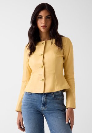 Femme aux cheveux foncés portant une veste jaune ajustée boutonnée et un jean bleu sur un fond clair uni.