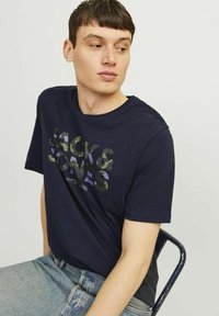 Tricou din bumbac bleumarin, cu un imprimeu de logo în camuflaj în nuanțe de verde și violet. Mâneci scurte și guler rotund. Purtați cu blugi deschiși la culoare.