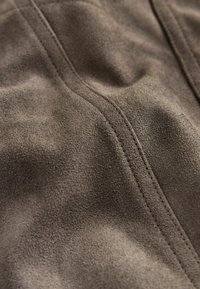 Bruin suède stof met een zachte textuur, met subtiele rimpels en stiksel details in een donkere draad, wat een glad uiterlijk creëert.