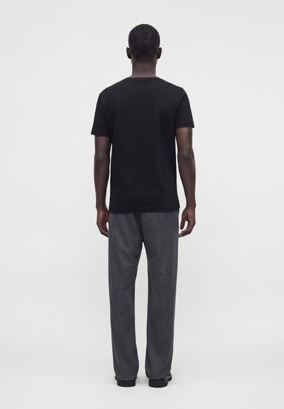 Homme debout de dos, portant une chemise noire à manches courtes et un pantalon gris ample sur un fond clair uni.