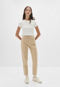 Witte aansluitende t-shirt gecombineerd met beige hoge taille jeans en lichtgekleurde sneakers. Model staat met handen op de heupen tegen een effen achtergrond.