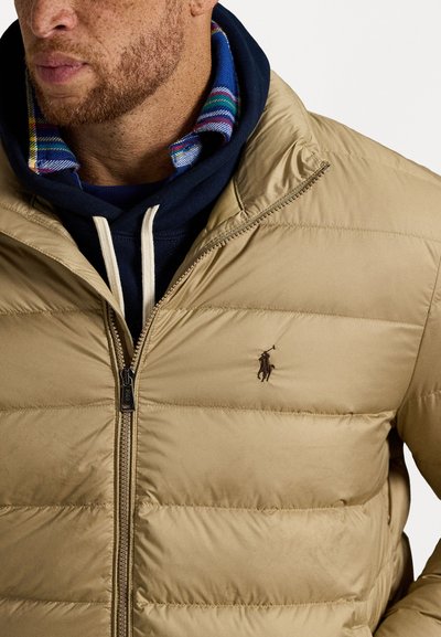 Beige pufferjakke med vandret stikninger, lynlåslukning og et lille broderet logo. Lagt over en blå hættetrøje og en multicolor krave.