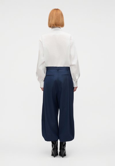 Femeie cu păr roșu scurt, purtând o bluză albă, pantaloni largi cropați cu dungi bleumarin și botine negre cu toc, stând cu fața spre spate.