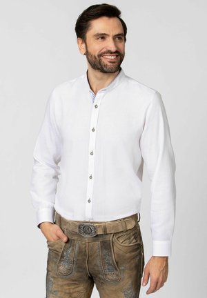 Weißes Langarm-Hemd mit Knopfleiste und Mandarin-Kragen, ausgestattet mit silbernen Knöpfen und einem dezenten blauen Muster im Innenfutter. Kombiniert mit bestickten braunen Hosen.
