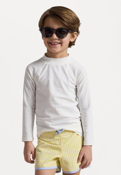 Rash guard blanca de manga larga combinada con pantalones cortos de baño a cuadros amarillos, con un cordón azul claro, llevada con gafas de sol oscuras.