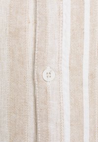 Tela con rayas en tonos marrón claro y crema, con un tejido texturizado y un detalle de botón blanco. Presenta líneas verticales de anchos variados.