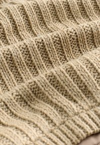 Beige gestrickte Decke mit dicken und dünnen Rippmustern, die einen weichen, strukturierten Look und leicht unregelmäßige Stiche für zusätzliche Tiefe aufweist.