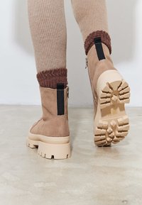 Tan suède enkellaarsjes met een chunky beige zool. Voorzien van een zwarte trekstrip, rits en een geribde bruine brei bovenaan. Getextureerde oppervlakte.