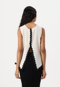 Haut sans manches beige avec des bordures festonnées noires et un détail en trou de serrure au dos, fabriqué dans un tissu texturé. Porté par-dessus une jupe noire ajustée.