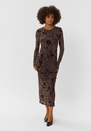 Femme aux cheveux bouclés portant une robe marron à manches longues avec des motifs floraux noirs et des talons hauts noirs, debout devant un fond uni.
