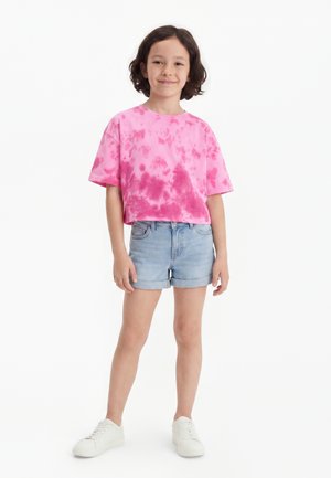 Enfant aux cheveux courts et foncés portant un t-shirt rose tie-dye, un short en jean clair et des baskets blanches, debout et souriant sur un fond blanc.