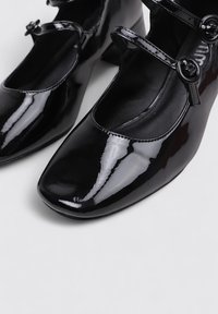 Chaussures Mary Jane en cuir verni noir avec un bout rond, des sangles à double boucle et un petit talon carré. Surface lisse et brillante.