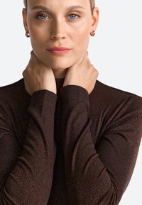 Braunes, geripptes Oberteil mit langen Ärmeln und subtilen Glitzereffekten; Model hält sich am Hals, trägt goldene Ohrringe und einen neutralen Lippenstift.