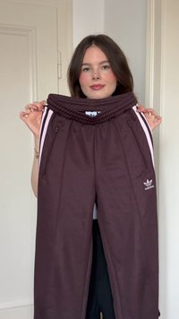Ruskeat urheilusortsit, joissa on teksturoitu, joustava vyötärö, vaaleanpunaiset sivusuorat, vetoketjulliset taskut ja etupuolella esillä oleva Adidas-logo.