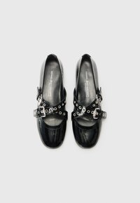 Black patent leather shoes with a rounded toe, featuring crisscross straps with grommets and buckles for adjustable fit.