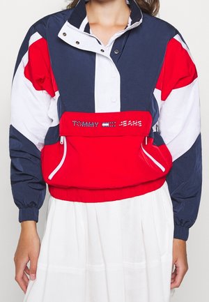 Vrouw draagt een rode, witte en marineblauwe Tommy Jeans windbreaker met voorzakken met rits en elastische manchetten, gecombineerd met een witte rok.