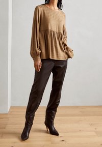 Kvinde iført en løs beige langærmet bluse og tætsiddende sorte læderbukser med høje hæle, stående på et trægulv.