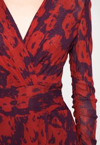 Robe à motifs rouges et violets avec un décolleté en V profond, des manches longues et des détails froncés au poignet, fabriquée en tissu texturé.