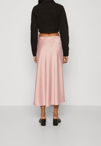 Woman standing, wearing a black cropped jacket, a pink satin midi skirt, and black heeled mules, against a plain light background.