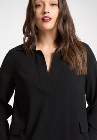 Tunica nera con colletto a scollo a tacca, maniche lunghe e tasche frontali, caratterizzata da una consistenza liscia e una vestibilità comoda.