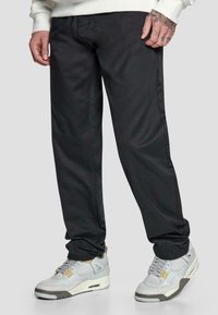 Schwarze, schmal zulaufende Hose aus glattem Stoff mit seitlichen Nähten, kombiniert mit grauen Sneakern mit gelben Akzenten und einem strukturierten Design.