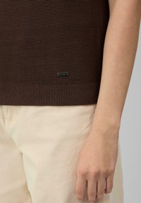 Pull en maille marron avec ourlet à côtes, arborant une petite étiquette logo noire. Porté par-dessus un pantalon beige clair, mettant en valeur la texture et le design décontracté.
