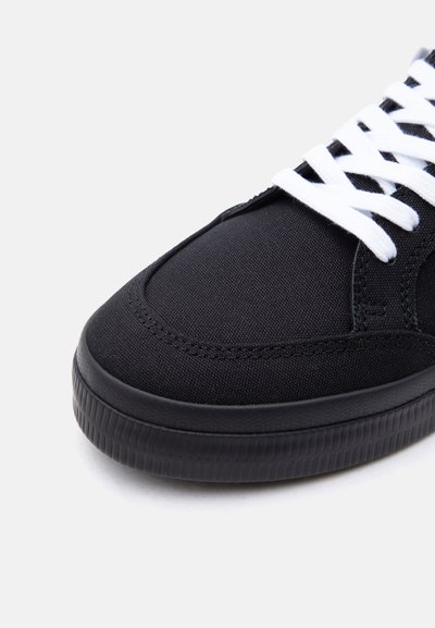 Κοντινό πλάνο μαύρου υφασμάτινου αθλητικού παπουτσιού με λευκά κορδόνια και υφή μαύρης λαστιχένιας σόλας σε λευκό φόντο.